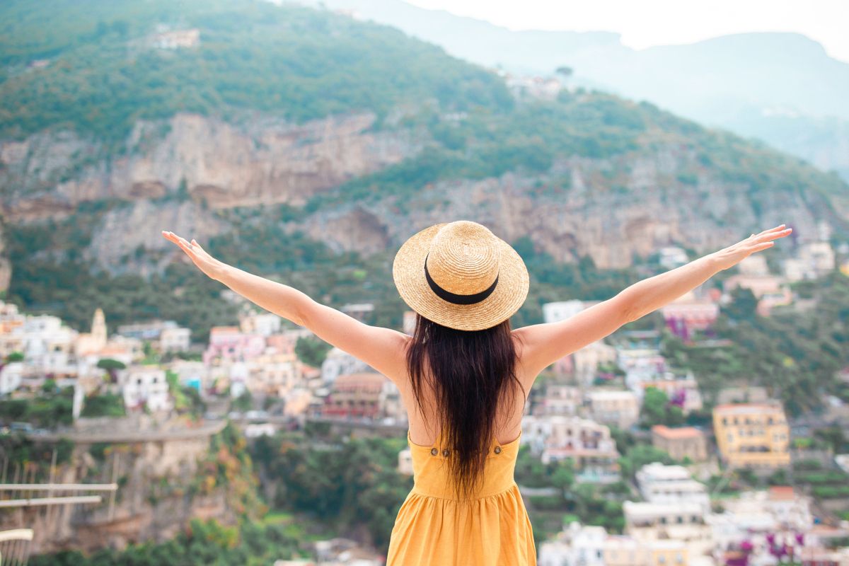 Back view of young woman in straw hat and yellow dress with Positano village on the background, Amalfi Coast, Italy