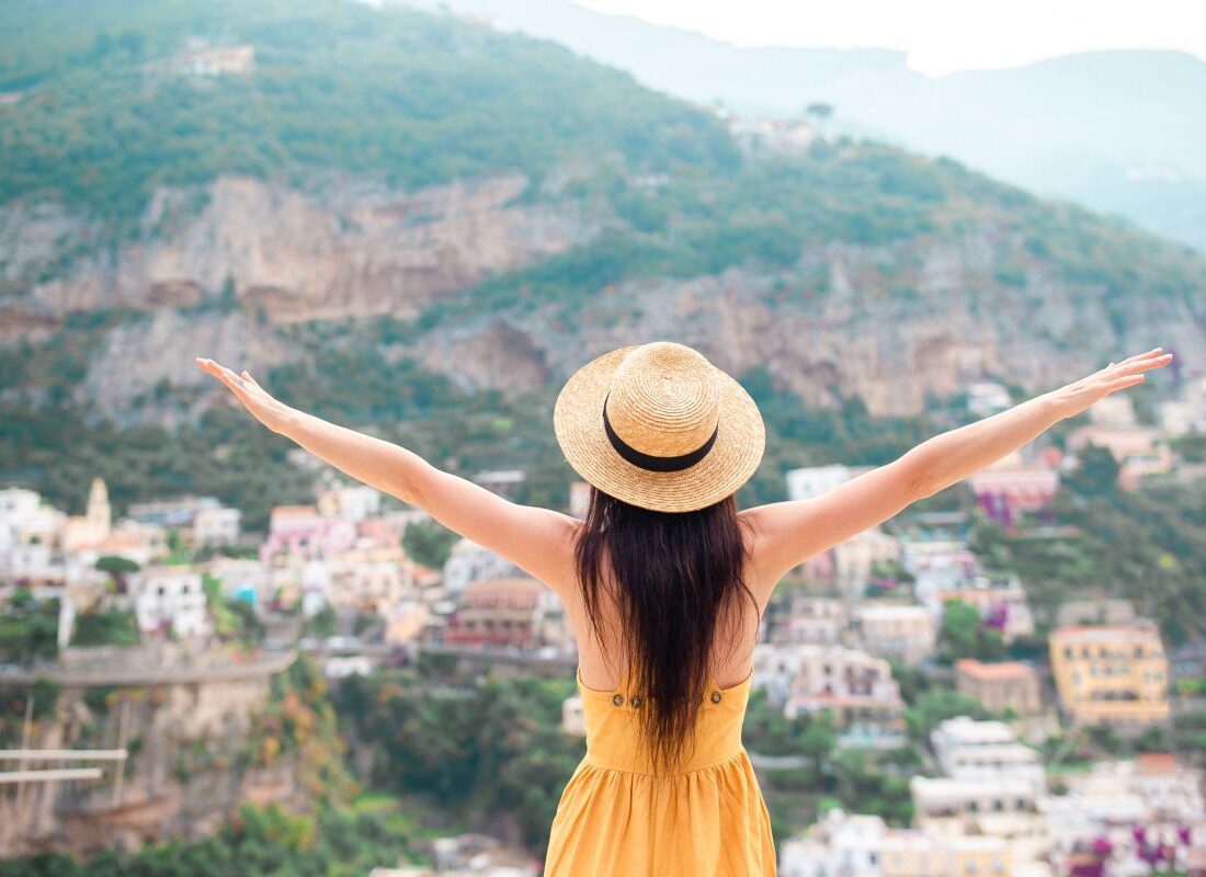 Back view of young woman in straw hat and yellow dress with Positano village on the background, Amalfi Coast, Italy