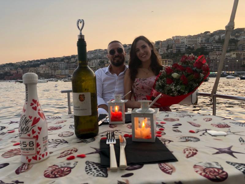 coppia in barca, lei con un mazzo di fiori, tavolo apparecchiato con bottiglia di vino, petali, calici, due candele all'interno di due lanterne